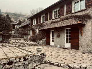 Maison rustique en pierre avec volets en bois, entourée de végétation et d'un chemin pavé, située dans un cadre montagnard