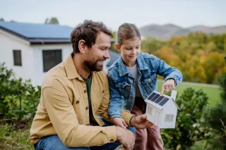 adobestock-540667187-1-webp.webp Un homme et une fille sourient en tenant une maquette de maison équipée de panneaux solaires, près d'une maison réelle avec des panneaux solaires