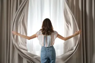 aeration_maison_fenetre.webp Une femme ouvre des rideaux beige pour laisser entrer la lumière du jour.