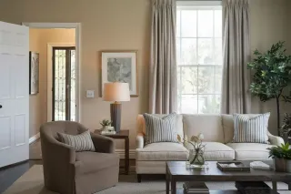 choisir_triple_vitrage_isolation.webp Un salon confortable avec un canapé beige, un fauteuil brun, une table basse, des coussins rayés et une grande fenêtre éclairant la pièce.