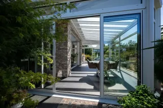 connexion_nature_veranda.webp Une véranda élégante aux murs en verre et pierre, baignée de lumière naturelle, donnant sur un jardin verdoyant.