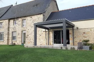difference_pergola_veranda.webp Une maison en pierre avec une pergola moderne en métal gris sur une pelouse verte.