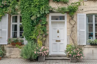 durabilite_vitrage.webp Façade charmante d'une maison en pierre avec une porte blanche entourée de vignes grimpantes et de fleurs colorées.