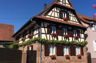 entretien_volets.webp Maison à colombages traditionnelle avec des fleurs rouges aux fenêtres, située sous un ciel bleu clair.