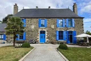fenetre_couleur_maison_tryba.webp Maison en pierre avec volets et porte bleus, entourée de verdure et d'un jardin aménagé, sous un ciel partiellement nuage