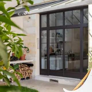 fenetre_croisillons.webp Une grande baie vitrée en métal noir s'ouvre sur une cuisine moderne, entourée de murs en pierre claire, avec des plantes et une pile