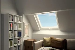 fenetre_de_toit_accessoire.webp Une pièce lumineuse sous les combles, aménagée avec une grande fenêtre de toit, une bibliothèque et un canapé confortable.