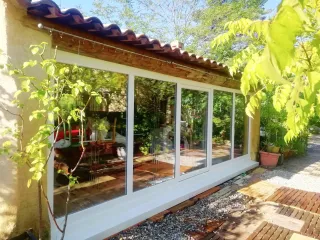 fenetre_fixe_pvc_tryba.webp Une véranda lumineuse avec de grandes baies vitrées entourée de verdure et de plantes en pots.