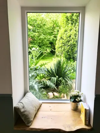 Fenetre_fixe_pvc.webp Un coin lecture cosy avec une fenêtre donnant sur un jardin verdoyant, un coussin, un livre et une plante en pot sur une étagère