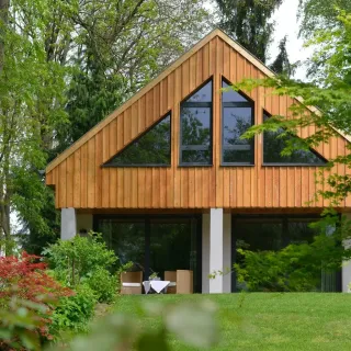 fenetre_formes.webp Maison moderne en bois avec de grandes fenêtres triangulaires, entourée de verdure luxuriante.