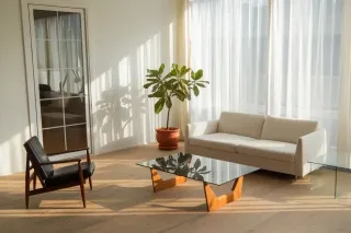 fenetre_salon_porte.webp Un salon lumineux et minimaliste avec un canapé blanc, une table basse en verre, un fauteuil en bois foncé et une plante en pot, ba