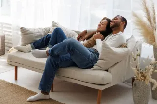 fenetre_securisee.webp Un couple souriant est assis confortablement sur un canapé beige dans un salon lumineux et décoré de plantes.