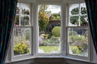 fenetre_securite_infraction.webp Vue d'un jardin serein à travers une fenêtre aux vitres brisées, encadrée de rideaux colorés.