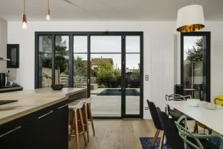 fenetre_vantaux_un_deux.webp Cuisine moderne et lumineuse avec vue sur une terrasse en bois et piscine, aménagée avec un îlot central et des chaises design.