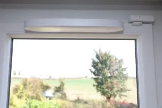 Grille_hygrorglable.webp Vue à travers une fenêtre avec un détecteur de contact installé en haut, donnant sur un paysage flou de verdure et un arbre.