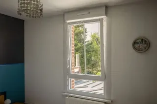 Grille_standard.webp Une fenêtre moderne s'ouvre sur un jardin verdoyant, éclairant une pièce aux murs blancs avec une horloge murale.