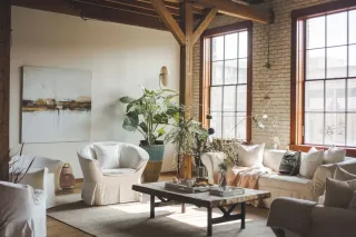 Indice_Rw.webp Un salon lumineux au style industriel avec mobilier blanc, grandes fenêtres, et plantes d'intérieur verdoyantes.
