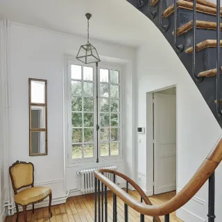 inspiration_fenetre_couleur.webp Un hall d'entrée lumineux avec un escalier élégant, une fenêtre à grandes vitres, un radiateur moderne et une chaise classique.