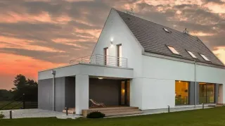 Isolation-by-tryba.webp Maison moderne blanche avec un toit gris, éclairée en soirée par un ciel orangé.