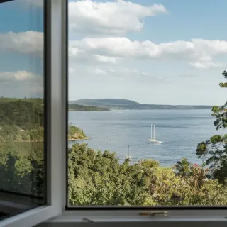 joints_mastics_fenetre.webp Vue pittoresque d'une fenêtre ouverte donnant sur un paysage côtier avec un voilier et une végétation verdoyante.