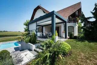 lames_orientable_pergola.webp Maison moderne avec terrasse couverte et jardin verdoyant, intégrant une piscine et une vue sur la campagne environnante.