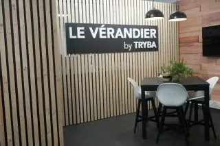 Le_verandier_by_tryba.webp Espace moderne avec un panneau 'LE VÉRANDIER by TRYBA', des chaises blanches autour d'une table noire, et une décoration