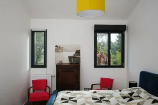 Motorisation_volets.webp Une chambre lumineuse avec deux fenêtres, des fauteuils rouges, un lit avec une couverture géométrique, et une décoration murale apaisante