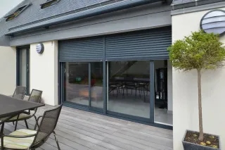 option_installation_volets_1.webp Façade de maison moderne avec grandes baies vitrées et volets roulants, donnant sur une terrasse en bois équipée de mobilier extérieur.
