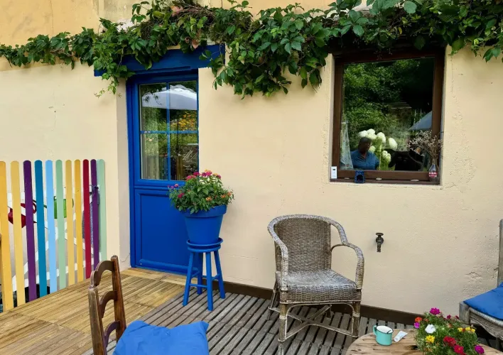 pe-tryba-pvc-bleu__1_.webp La terrasse colorée et fleurie comporte des chaises en osier et une porte bleue, offrant un espace convivial et accueillant.