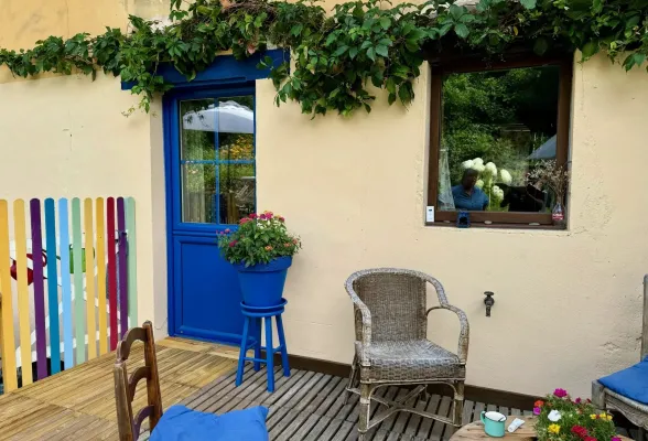 pe-tryba-pvc-bleu__1_.webp La terrasse colorée et fleurie comporte des chaises en osier et une porte bleue, offrant un espace convivial et accueillant.