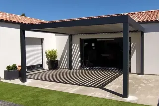 pergola_aluminium.webp Une maison moderne avec pergola en métal noir surplombant une terrasse partiellement ombragée, entourée de murs blancs et de tuil