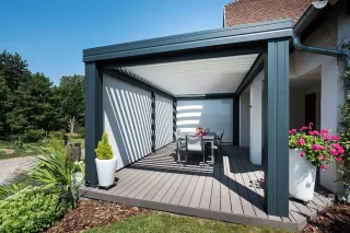 pergola_bioclimatique_consommation_energetique.webp Une élégante pergola moderne en métal gris abrite une terrasse bois avec une table à manger entourée de plantes fleuries.