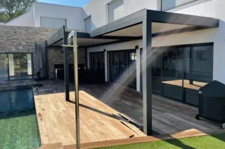 pergola_bioclimatique_tryba.webp Terrasse moderne avec pergola en métal et piscine, donnant sur une maison contemporaine aux murs en pierre.
