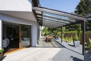 pergola_volutive_a_toit_fixe.webp Terrasse moderne avec pergola en verre, mobilier de jardin en bois, et plantes luxuriantes, idéale pour profiter de l'extérieur.
