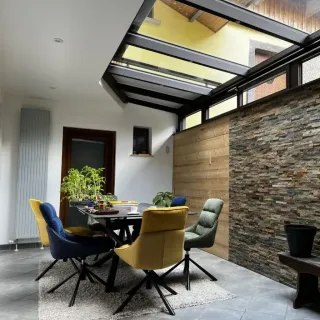 polyvalence_veranda.webp Salle à manger moderne avec toit en verre, murs en pierre et bois, et chaises colorées autour d'une table.