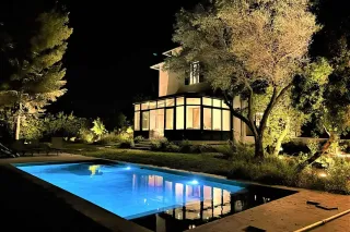 premontage_usine_veranda.webp Une maison moderne illuminée de nuit avec une piscine scintillante et un jardin verdoyant.