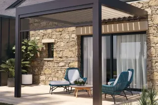 protection_pergola_tryba.webp Une terrasse élégante avec des fauteuils en osier bleu sous une pergola moderne adossée à un mur en pierre.