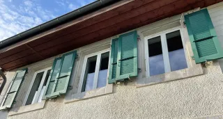 pvc.webp Façade d'une maison avec des volets verts en bois ouverts sur des fenêtres blanches, sous un toit en bois.