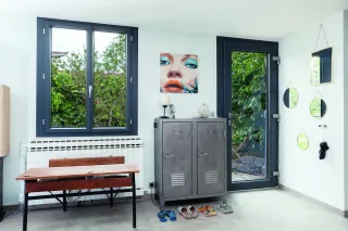 Realisation_ALU_gris_anthracite_TRYBA_bordeaux__2_.webp Un intérieur lumineux avec de grandes fenêtres, une armoire métallique, des tableaux et des miroirs muraux, et des claquettes posées au sol