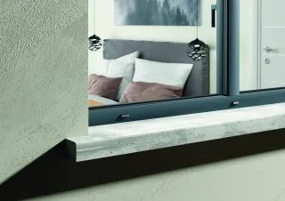 TA76oc_DETAIL_DORMANT.webp Vue à travers une fenêtre moderne avec rebord en béton, montrant une chambre contemporaine avec un lit à coussins mauves et des suspensions