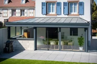 technologie_3D_veranda.webp Une maison moderne avec une véranda en métal gris dotée de grandes baies vitrées, donnant sur une terrasse en dalles claires et un