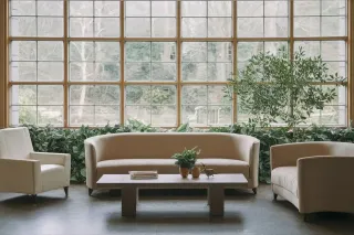 triple_vitrage_factures_energies.webp Un salon lumineux avec des fauteuils crème, une table basse orné de plantes vertes en pot, et une grande fenêtre offrant une vue sur le
