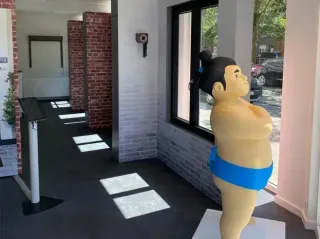 tryba_belgique_Magasin_Herentals.webp Une statuette colorée de sumo en tenue traditionnelle bleue se tient près d'une fenêtre dans une pièce aux murs de briques et carrel
