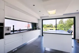 TRYBA_coulissant_aluminium_gris_anthracite.webp Cuisine moderne et lumineuse avec des éléments blancs, un plan de travail noir et de grandes baies vitrées ouvrant sur un jardin verdoyant.