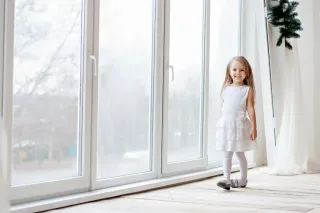 tryba_solution_sur_mesure_ventilation.webp Une petite fille souriante en robe blanche se tient près d'une grande baie vitrée.