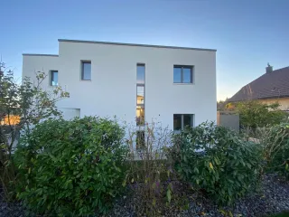 tryba_suisse_maison_fenetre.webp Façade moderne d'une maison blanche entourée de verdure par une journée ensoleillée.