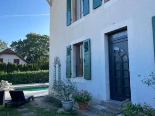 tryba_suisse_maison_porte_entree.webp Façade d'une maison avec volets verts, porte d'entrée sombre et piscine extérieure entourée de verdure.