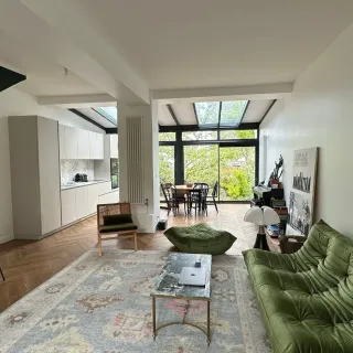 veranda_ou_extension_maison.webp Un salon moderne et lumineux avec un canapé vert, une table basse en marbre, et une grande baie vitrée donnant sur une terrasse.