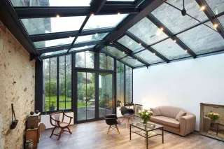veranda_qualite_materiaux.webp Une véranda moderne avec un toit en verre, un mobilier contemporain, et une vue sur un jardin verdoyant.
