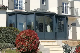 veranda_surface_habitable.webp Une véranda moderne en verre et métal gris anthracite jouxte une maison avec des volets gris, entourée de buissons soigneusement